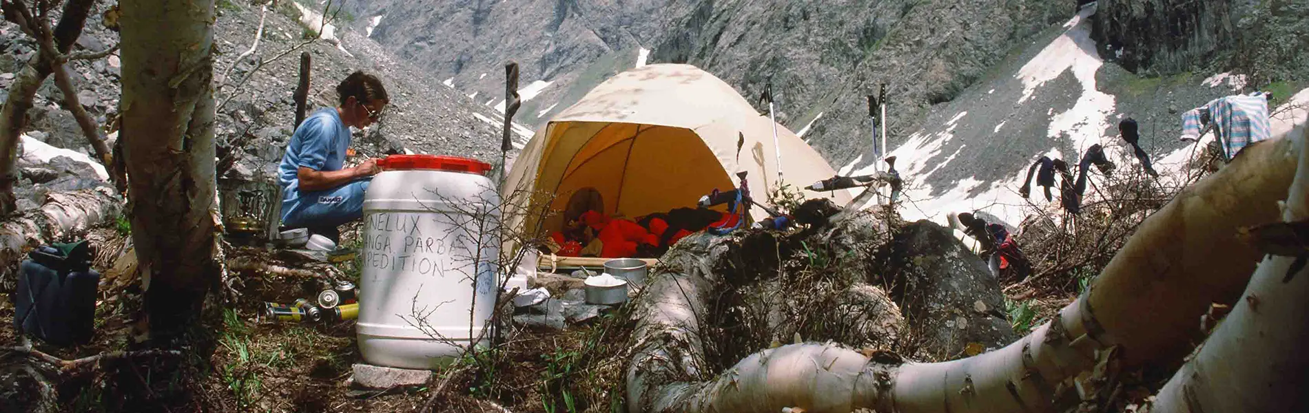 Twee bergbeklimmers hebben hun kamp opgeslagen op een besneeuwde berghelling, vlakbij een gele tent, met rondom verspreid liggende bergbeklimmersuitrusting. Op de achtergrond rijst onder een heldere h